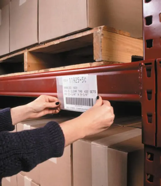 Hands placing a plastic label holder on a warehouse shelf with boxes and a wooden pallet above.