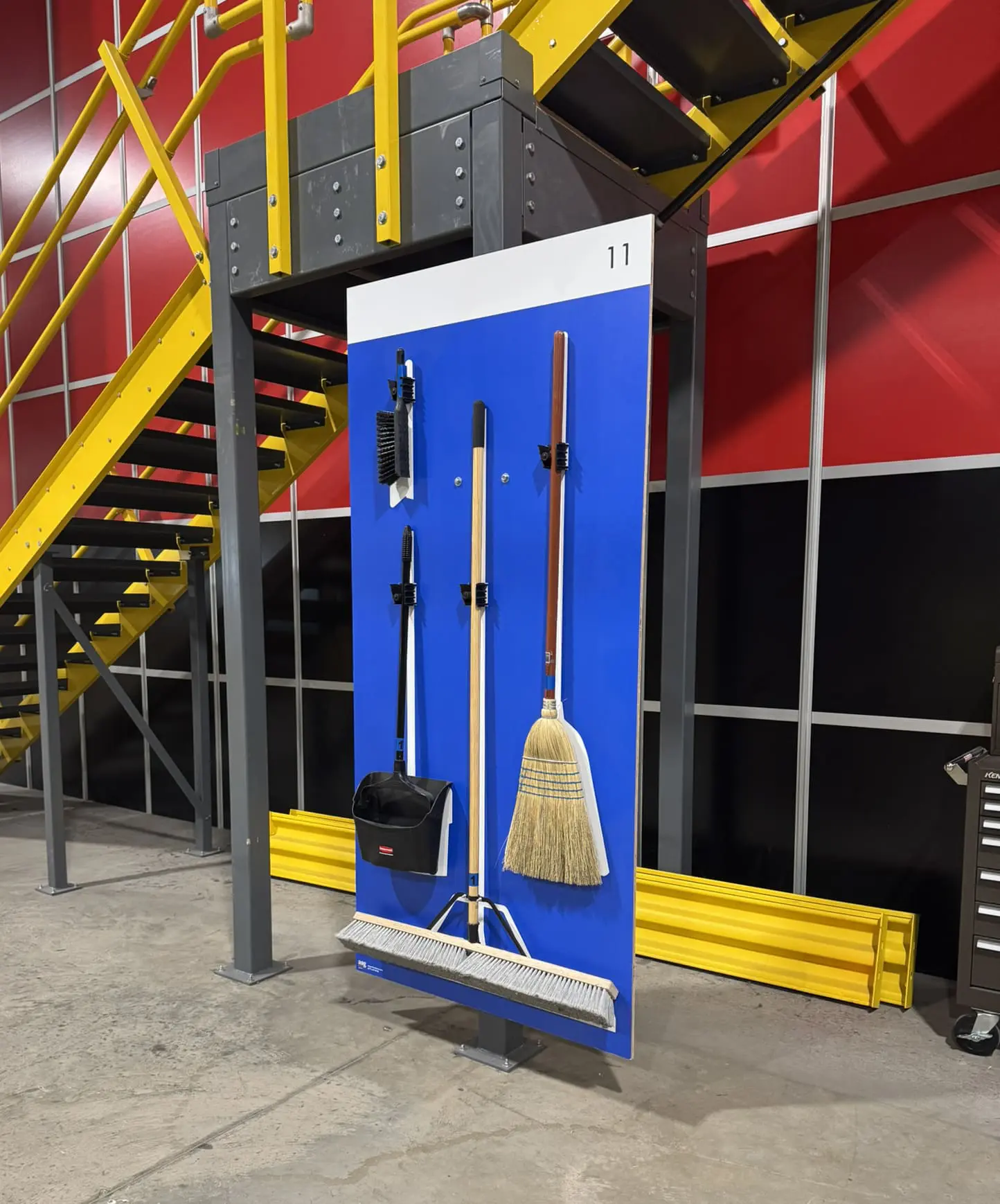 Cleaning tools hang on a blue board near yellow stairs in an industrial setting.