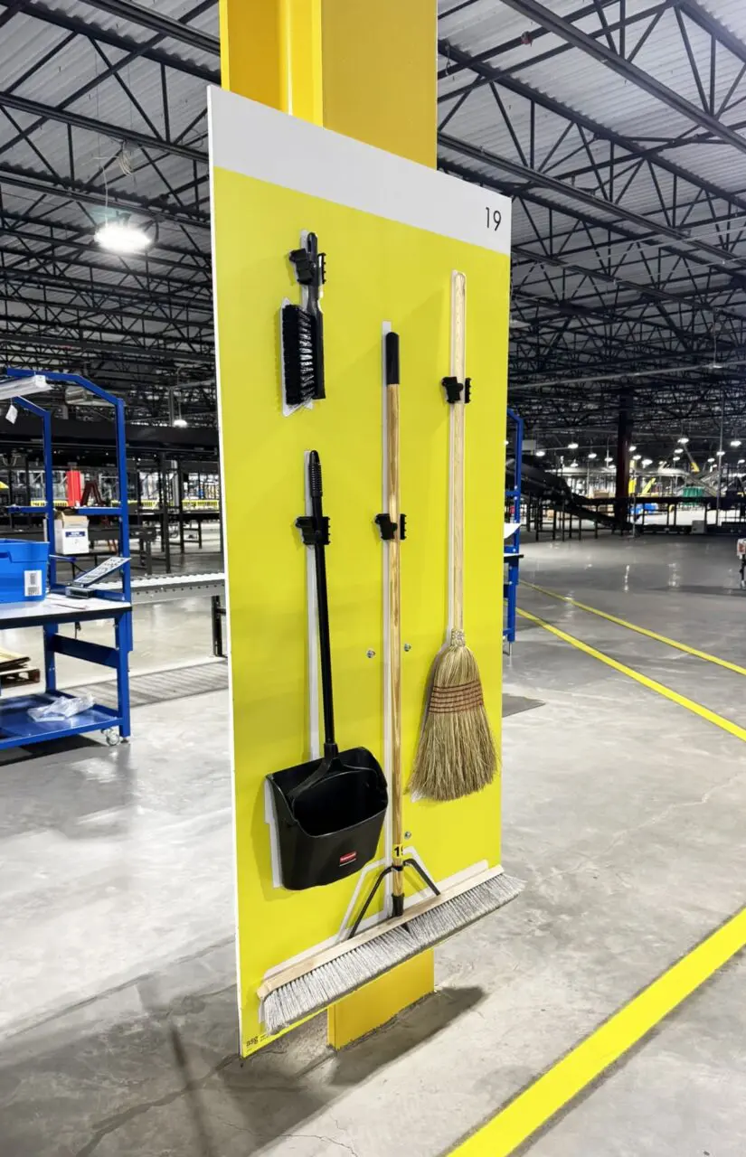 Wall-mounted cleaning tools: a broom, dustpan set, scrubbing brush, and mop in a warehouse setting.